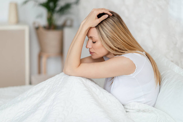 Wanhoop ongelukkige jonge blonde Europese vrouw die lijden aan slapeloosheid en hoofdpijn zitten op bed - Foto, afbeelding