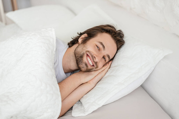Χαμογελώντας χιλιετή καυκάσιος άνδρας με γένια ξυπνήσει, βρίσκεται στο λευκό κρεβάτι, απολαμβάνει ελεύθερο χρόνο και το Σαββατοκύριακο - Φωτογραφία, εικόνα