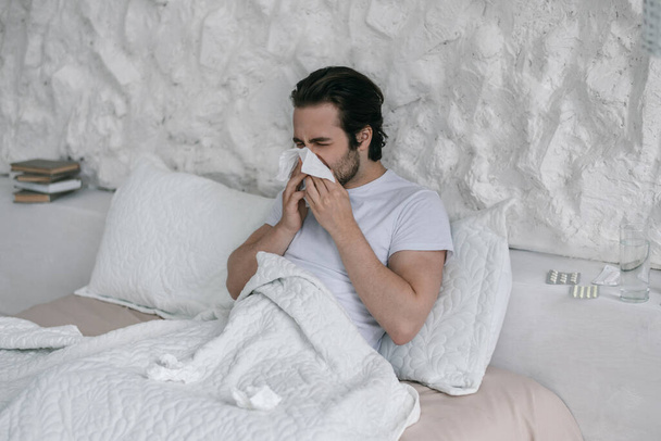 Unhappy millennial european male lies on bed, blows nose at napkin in white bedroom interior - Zdjęcie, obraz