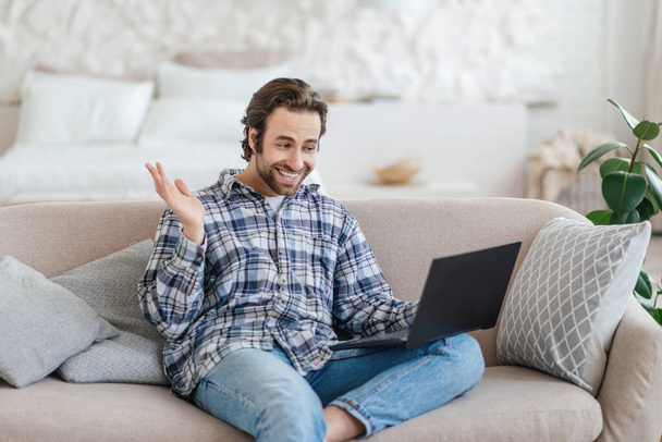 Satisfied millennial caucasian man with stubble sits on sofa waving hand and looking at laptop - 写真・画像