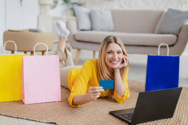 Tevreden jonge blonde Europese dame met laptop en creditcard ligt op de vloer met vele pakketten - Foto, afbeelding