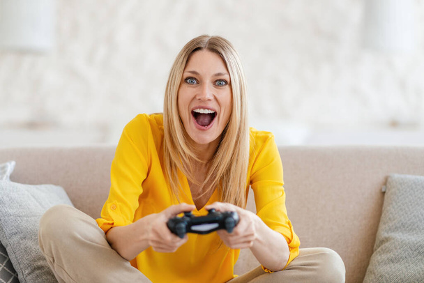 Взволнованная молодая блондинка в желтой одежде с открытым ртом играет в онлайн-игры - Фото, изображение