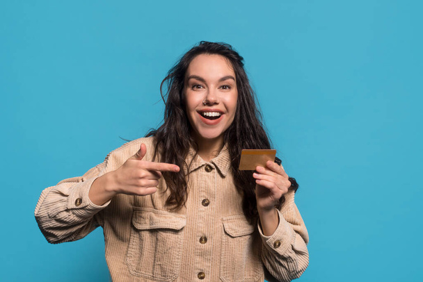 Весело удивлена юная белая брюнетка с открытым ртом указывает пальцем на кредитную карту - Фото, изображение
