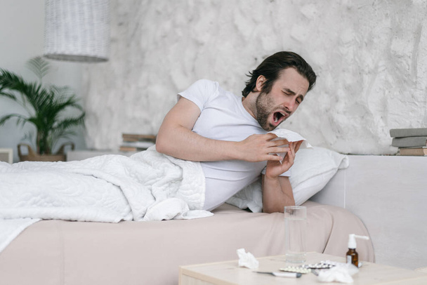 Sad millennial caucasian guy with stubble lies on bed, sneezes at napkin in bedroom interior with medicines - Fotó, kép