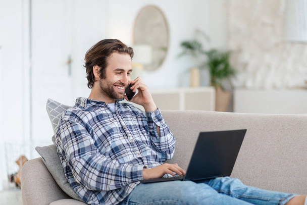 Wesoły, millenium biały mężczyzna ze ścierniskiem siedzi na kanapie, pisząc na laptopie i dzwoniąc przez telefon - Zdjęcie, obraz