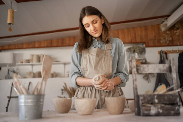 Young female artisan concentrated on modeling jug from raw clay while pottery lesson or workshop in creative studio. Woman ceramic business owner making craft for sale in handmade potter retail store - Fotó, kép