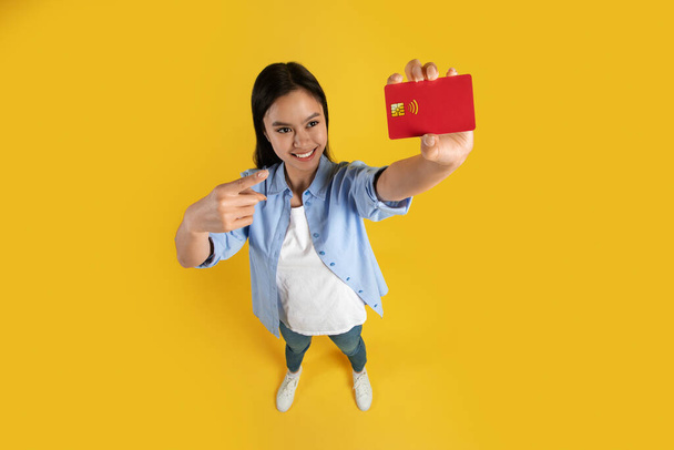 Feliz bastante divertido joven japonesa mujer en casual señalar el dedo en la tarjeta de crédito con espacio en blanco - Foto, imagen