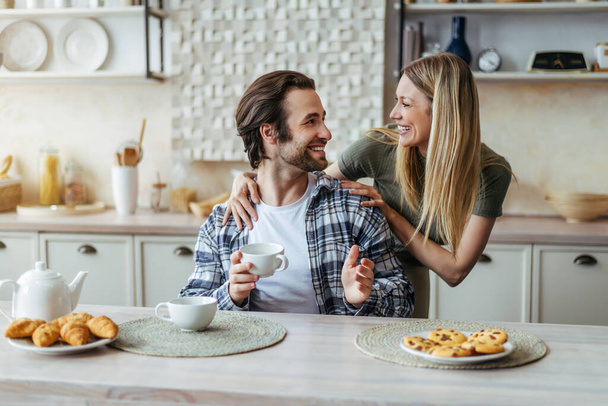 Cheerful young caucasian wife look at husband, drinking coffee and talking in free time in modern kitchen interior - Foto, imagen
