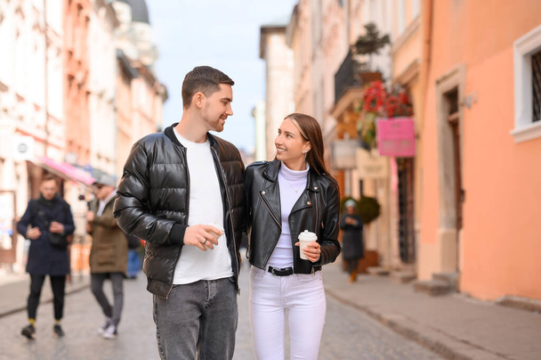 Preciosa pareja joven con tazas de café caminando juntos en la calle de la ciudad. Fecha romántica - Foto, imagen