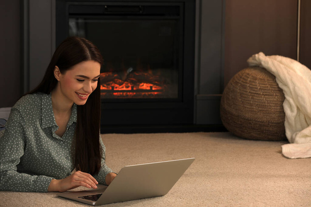 Beautiful young woman with laptop lying on floor near fireplace at home. Space for text - 写真・画像