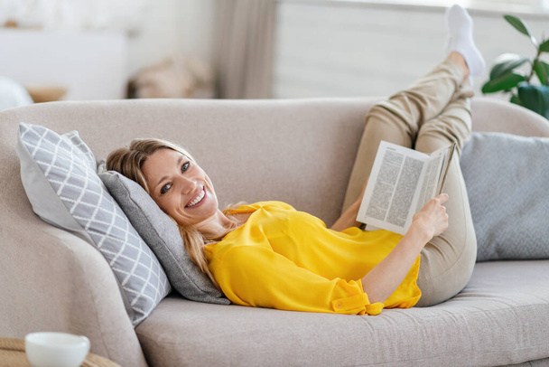 Smiling attractive millennial blond european lady in yellow clothes lies on sofa and reads book in living room - Fotó, kép
