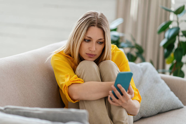 Sad young blonde european woman looks at phone, hugs her knees on sofa in living room interior - Zdjęcie, obraz