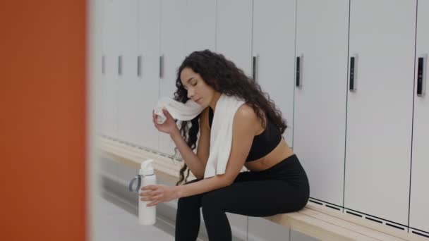Молодая активная спортсменка отдыхает после тяжелой тренировки в раздевалке спортзала, пьет воду и вытирает пот с лица - Кадры, видео