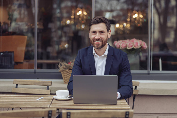 コーヒーショップのテラスにノートパソコンと笑顔で座っている白人ハンサムなエレガントなビジネスマン. - 写真・画像