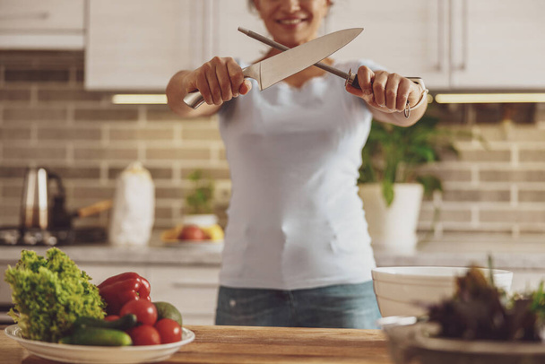 A housewife sharpens a knife standing at a table against the background of the kitchen - Фото, изображение