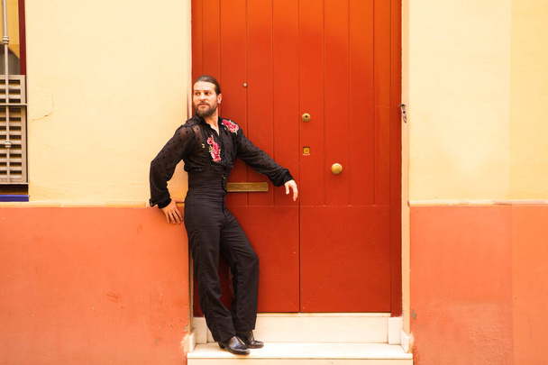 Mann tanzt Flamenco mit schwarzem Hemd und roten Rosen, auf dem Hintergrund einer roten Holztür wartet er angelehnt an die Tür. Flamenco-Tanzkonzept kulturelles Erbe der Menschheit. - Foto, Bild