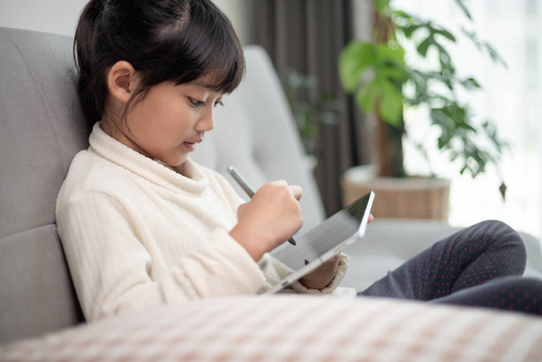 Malá dívka pomocí tabletu hraní hry na internetu, Kid sedí na pohovce sledování nebo mluvit s přítelem on-line, Dětská relaxace v obývacím pokoji v dopoledních hodinách, Děti s novou technologií koncept - Fotografie, Obrázek