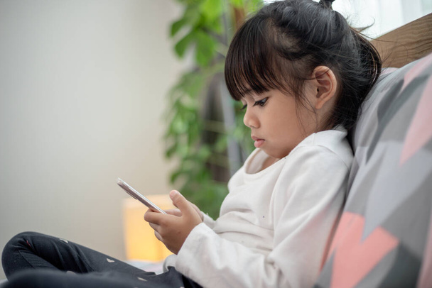 Cute little kid girl watching video on smartphone with smiley face alone on the bed, child using mobile phone with happy face at home. Stay at home quarantine coronavirus COVID-19 pandemic prevention. - 写真・画像