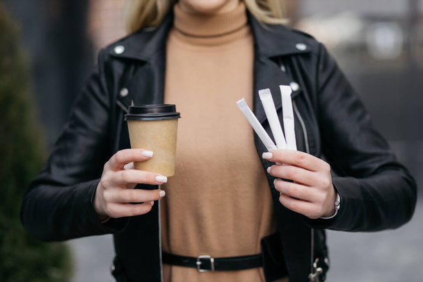Girl holding coffee in a paper cup and sticks with sugar on her face. Hands of a girl holding hot coffee on the background of the mall. - Fotoğraf, Görsel
