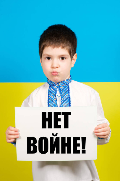 ロシア語で書かれた紙を持っているウクライナの民族衣装の子供はロシア語から翻訳-戦争はありません. - 写真・画像