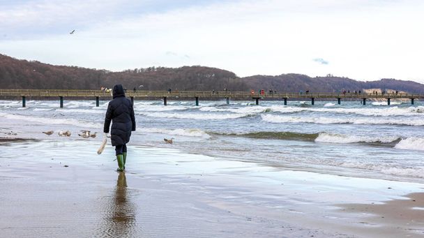 Een vrouw in een winterjas en laarzen loopt langs de kust. - Foto, afbeelding