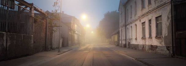 Route asphaltée éclairée près des bâtiments industriels modernes dans un brouillard. Des lanternes. Paysage urbain. Scène urbaine sombre. Industrie, infrastructures, pollution, économie, concepts écologiques - Photo, image