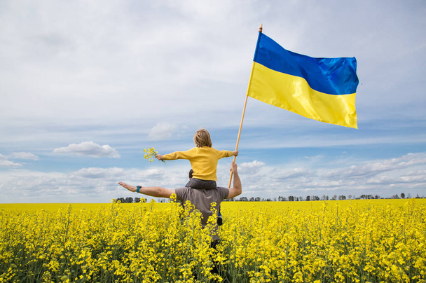 niño está sentado sobre los hombros de su padre, sosteniendo una gran bandera de Ucrania en las manos, de pie entre el campo de colza amarilla. educación del patriotismo. Orgullo, libertad. Ucranianos contra la guerra - Foto, Imagen