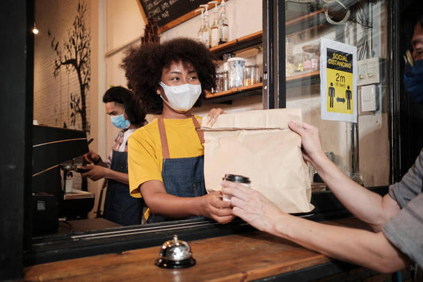 African American female barista with face mask works in the cafe with social distance, takeaway coffee for a customer, new normal service of small business coffee shop in COVID19 quarantine lifestyle. - Fotó, kép