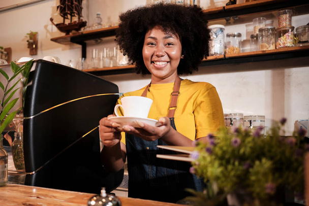 Afroamerykanka barista w spojrzeniu na aparat fotograficzny, oferuje filiżankę kawy do klienta z wesołym uśmiechem, happy service działa w swobodnej restauracji kawiarni, młody mały przedsiębiorca start-up. - Zdjęcie, obraz