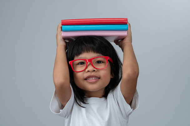 Funny and Happy Asian little preschool girl wearing red glasses holding a green book on the head, on white isolated background. Concept of school kid and education in elementary and preschool, home school - Foto, Bild