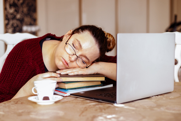 Mujer joven cansada en gafas durmiendo en libros cerca de la computadora portátil en el escritorio en la habitación de casa. Cansancio del estudio monótono y descanso en el lugar de trabajo del exceso de trabajo por computadora. Falta de concepto de motivación energética - Foto, imagen