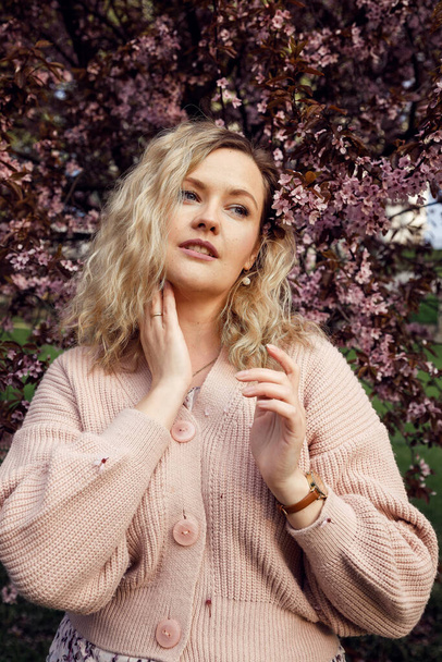 Portrait of beautiful blonde hair woman with gentle makeup and perfect curls posing on blooming lilac background in pink knitted cardigan. Fashion outfit for street walking on spring city park - Photo, Image