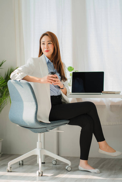 Young Asian businesswoman sitting at work holding coffee cup and facing back and laptop computer on financial business concept table - Foto, imagen