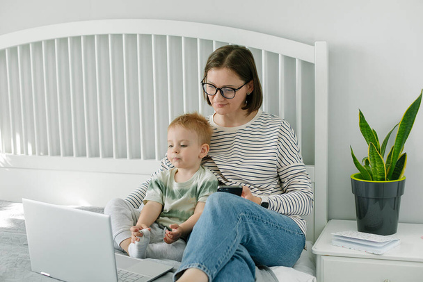 Young single mother mom with toddler boy son sitting on bed using laptop, searching web, watching cartoons together. Concept of work from home, freelancer - Fotó, kép