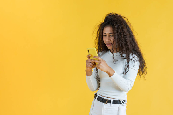 Göndör hajú fiatal nő használja a mobiltelefon sárga háttér - Fotó, kép