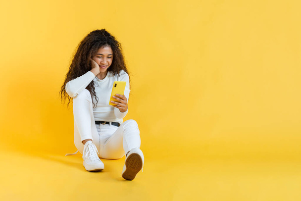 Young woman sitting using her phone entertaining herself against yellow background - Photo, Image