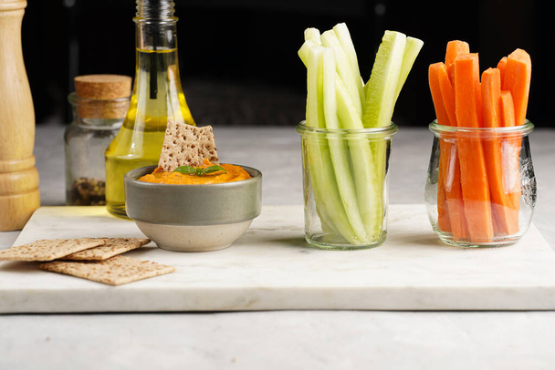 Paprika bell-pepper hummus dip looking like orange pumpkin puree, thin crackers with sesame seeds and carrot and cucumber cut in long strips and sticks on marble board - Fotoğraf, Görsel