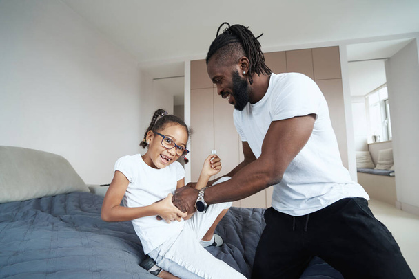 Adult African American man is tickling little daughter on bed - Photo, Image