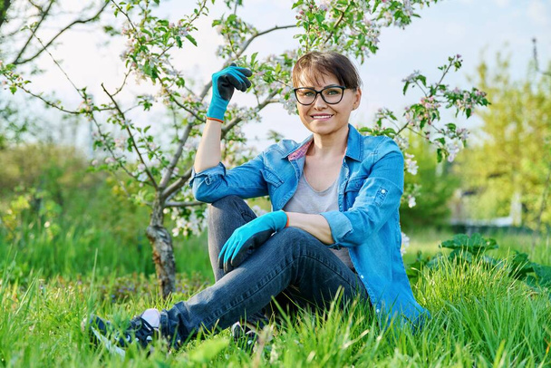 Positive beautiful mature female gardener sitting on the grass with blossoming apple tree. - Foto, afbeelding