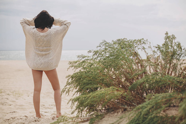 Piękna kobieta stojąca na piaszczystej plaży na tle zielonej trawy i patrząca na morze, spokojna, spokojna chwila. Stylowa młoda kobieta w beżowym swetrze z dzianiny relaksująca się na wietrznym zimnym wybrzeżu - Zdjęcie, obraz