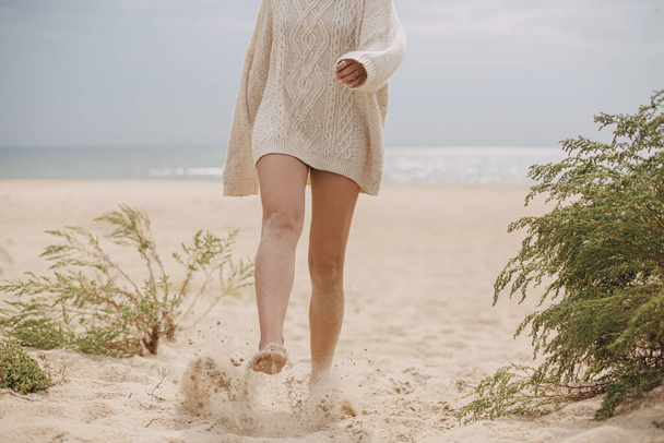 Beautiful woman legs barefoot with sand walking on sandy beach, carefree vacation mood. Stylish young female in knitted sweater relaxing on coast and having fun, cropped view - Photo, Image