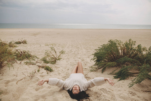 Schöne Frau liegt am Sandstrand vor dem Hintergrund von grünem Gras und Meer, unbeschwerter ruhiger Moment. Blick nach oben. Stylische junge Frau im Strickpullover genießt Urlaub und Entspannung an der Küste - Foto, Bild