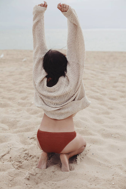 Hermosa mujer feliz con el pelo ventoso sentado en la playa de arena en el mar, momento sin preocupaciones. Elegante joven hembra en suéter de punto y bikini disfrutando de vacaciones y relajarse. Vista trasera. Imagen vertical - Foto, Imagen