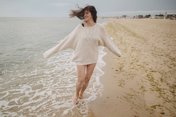 Mujer hermosa sin preocupaciones con el pelo ventoso corriendo en la playa de arena en las olas frías del mar, divirtiéndose. Elegante joven feliz hembra en suéter de punto relajante y disfrutar de vacaciones en la costa - Foto, Imagen