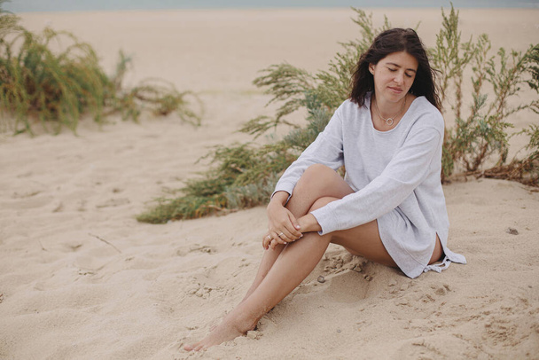 Schöne Frau mit windigen Haaren sitzt am Sandstrand vor dem Hintergrund von grünem Gras und Meer, ruhigen ruhigen Moment. Stylische junge Frau im Pullover entspannt an der Küste - Foto, Bild