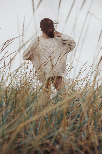 Gyönyörű, elegáns nő, szeles hajjal, kötött pulóverben, vad fű között pózolva. Gondtalan pillanat, stílusos kép. Divatos fiatal nő áll a szeles parton. Hiteles és nyugodt - Fotó, kép