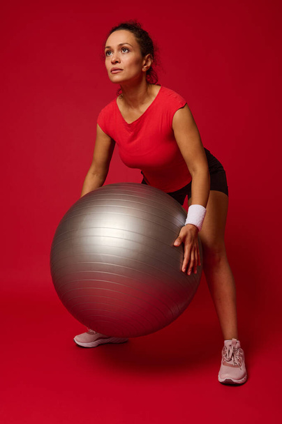 Full length portrait of a confident determined middle aged Hispanic sporty woman exercising with fitness ball against red background with copy ad space for advertising text - Φωτογραφία, εικόνα