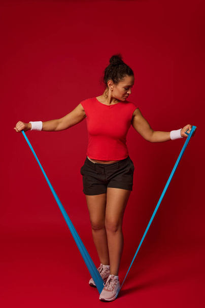 Full body size portrait of a sporty woman exercising with elastic fitness band while performing functional body weight training against res colored background with copy space. Fitness, sport concept - Photo, Image
