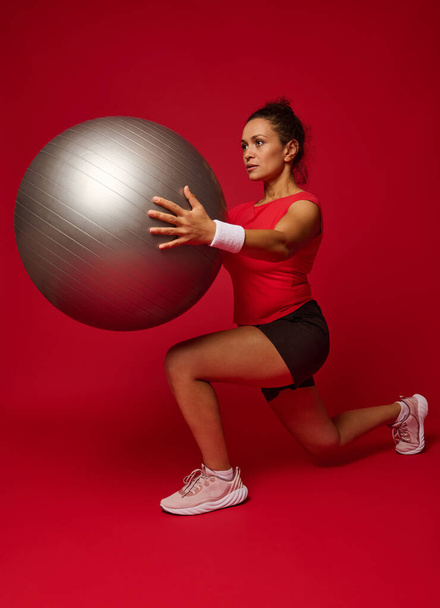 Attractive multi-ethnic young athletic woman doing forward lunges while exercising with a fit ball, holding it at chest level, isolated over red background with copy ad space. Sport, fitness concept - Zdjęcie, obraz