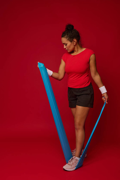 Full length of multi-ethnic sporty woman using elastic fitness rubber band exercises against red background with copy space for advertisement text. Fitness, sport, active and healthy lifestyle concept - Foto, Imagem